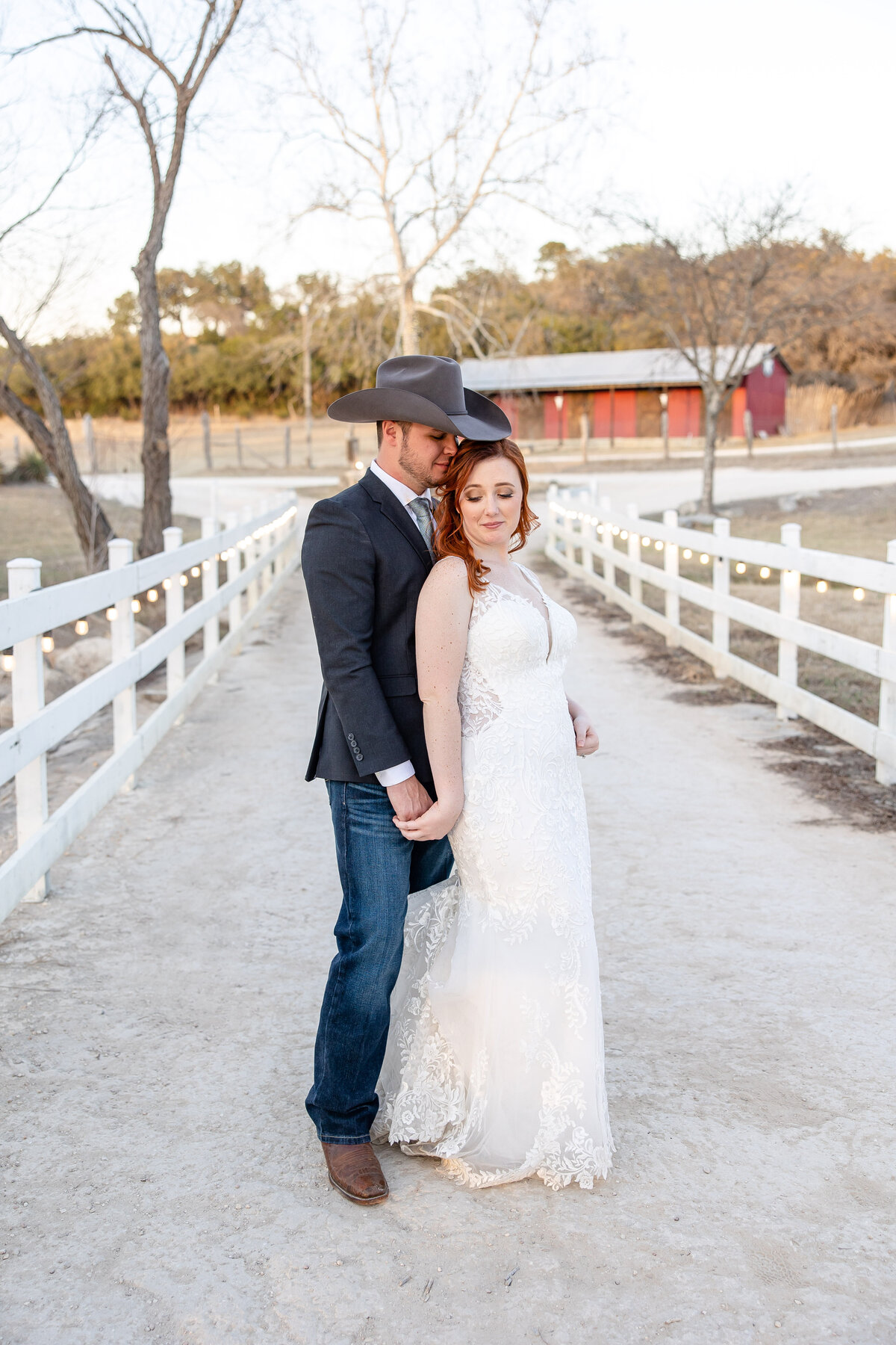 groom with cowboy hat stands with red head bride between white fences at Boerne Texas wedding