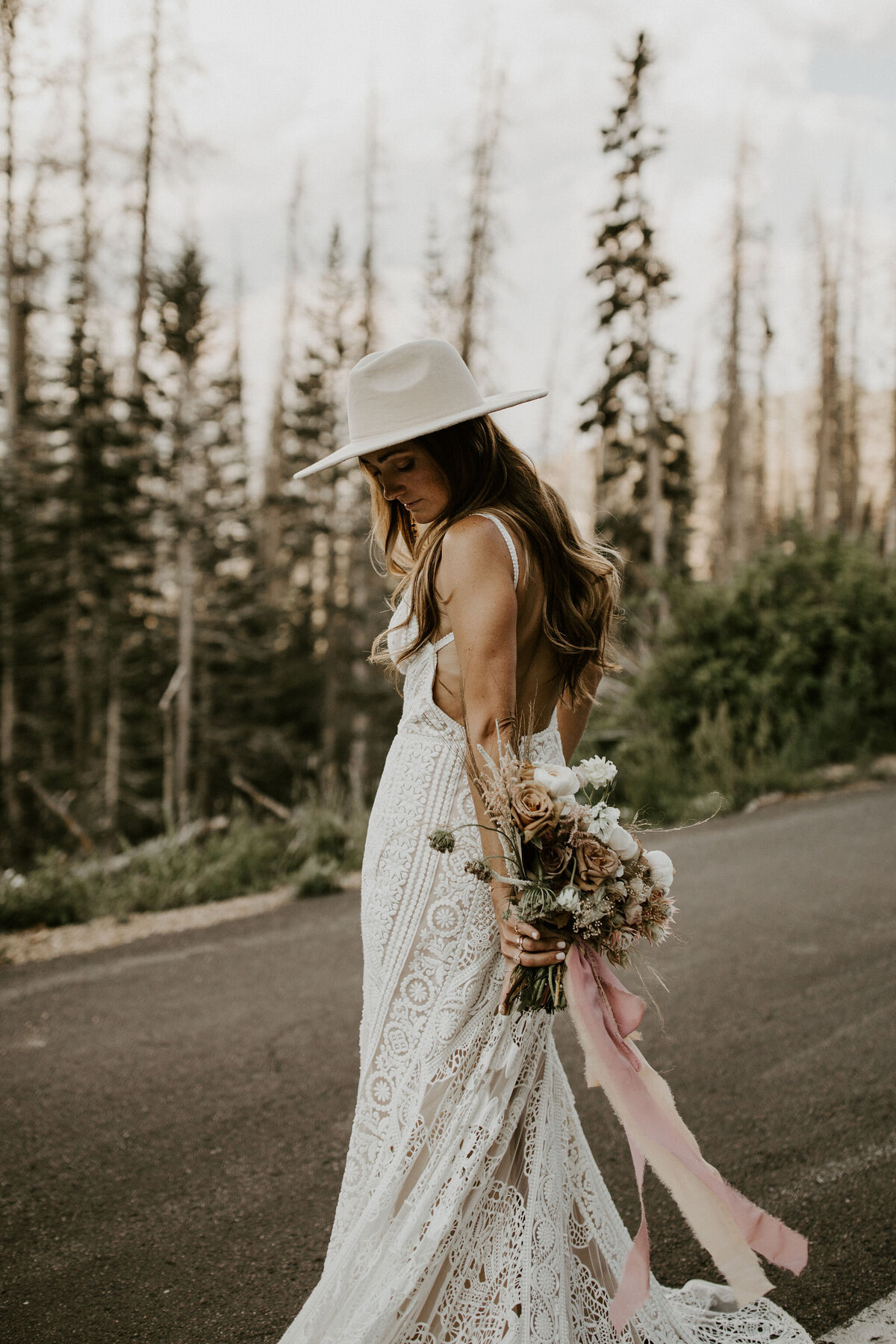 A bride wearing a white wedding gown and hat, holding a bouquet of blush and white roses on a road with trees in the background.