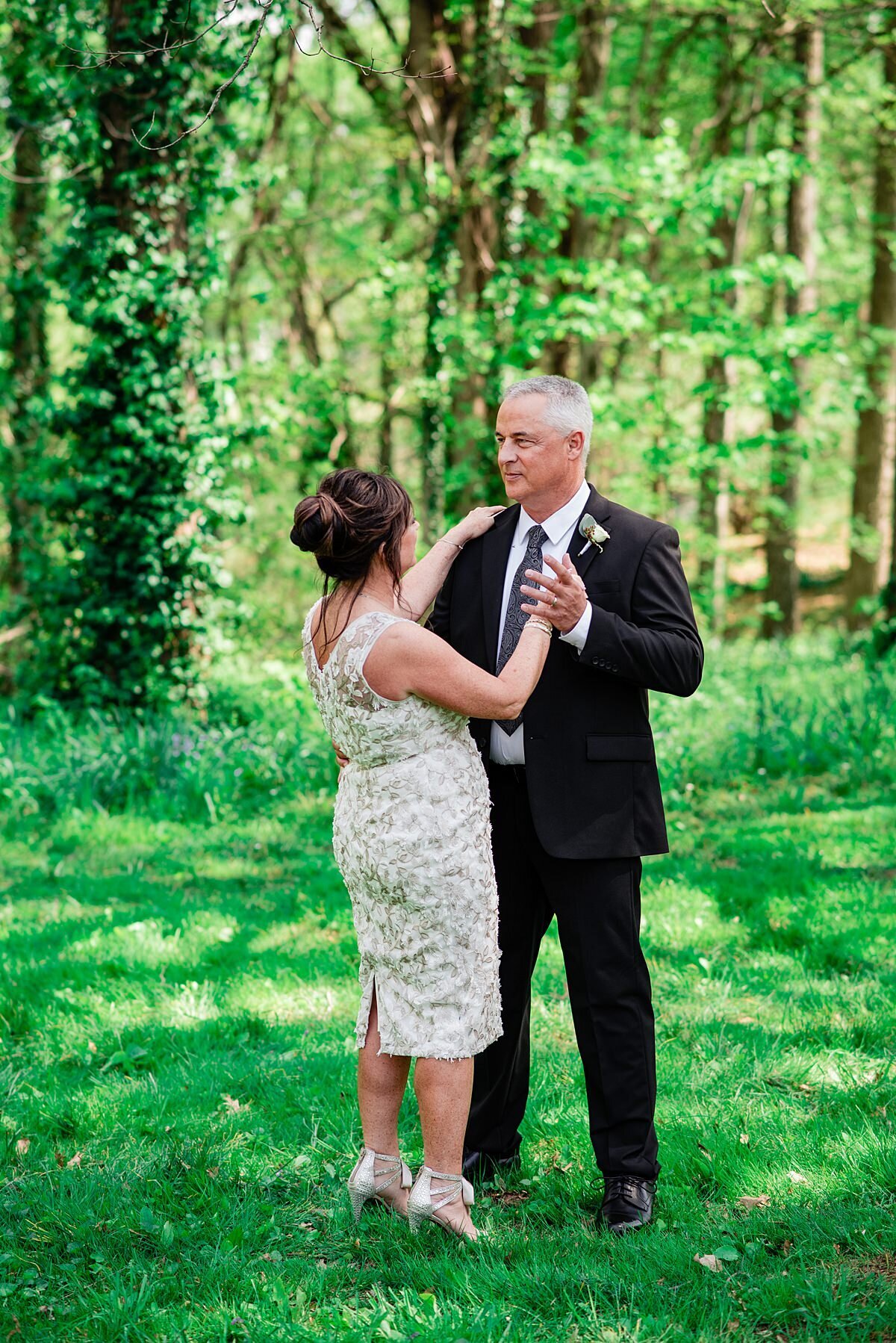 the bride and groom dance in the grass at the edge of the woods at Oaklands Mansion. The bride is wearing a tea length sheath sleeveless lace dress and stillettos with ankle bows. The groom is wearing a black suit with a dark patterned tie and a white boutonniere pinned to his lapel.