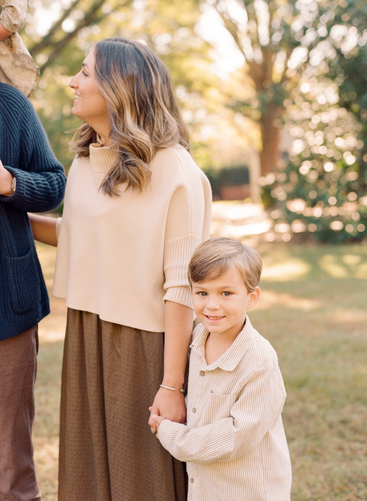 Son holds his mom's hands and smiles at the camera during their Raleigh family photography session. Family walking during their family portrait session in Wake Forest, NC. Photographed by Raleigh family photographer A.J. Dunlap Photography.
