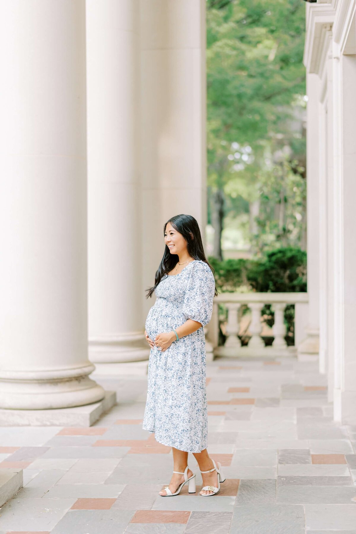 An expectant mother wearing a blue floral dress holds her growing belly while gazing off to the distance  through stately columns.  Photograph captured by Charlotte Maternity Photographer Melissa Mayrie Photography at Davidson College.