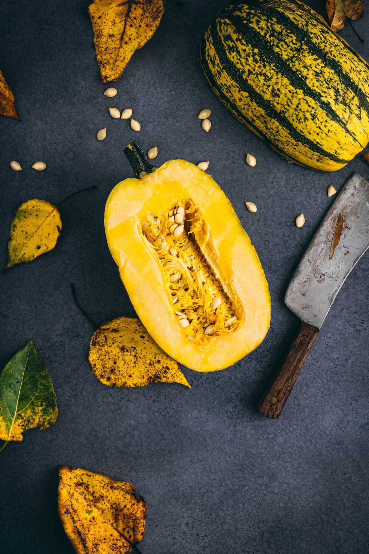 Fall leaves on dark grey surface with spaghetti squash cut in half with cleaver.  Leaves turning yellow and squash seeds scattered on surface.
