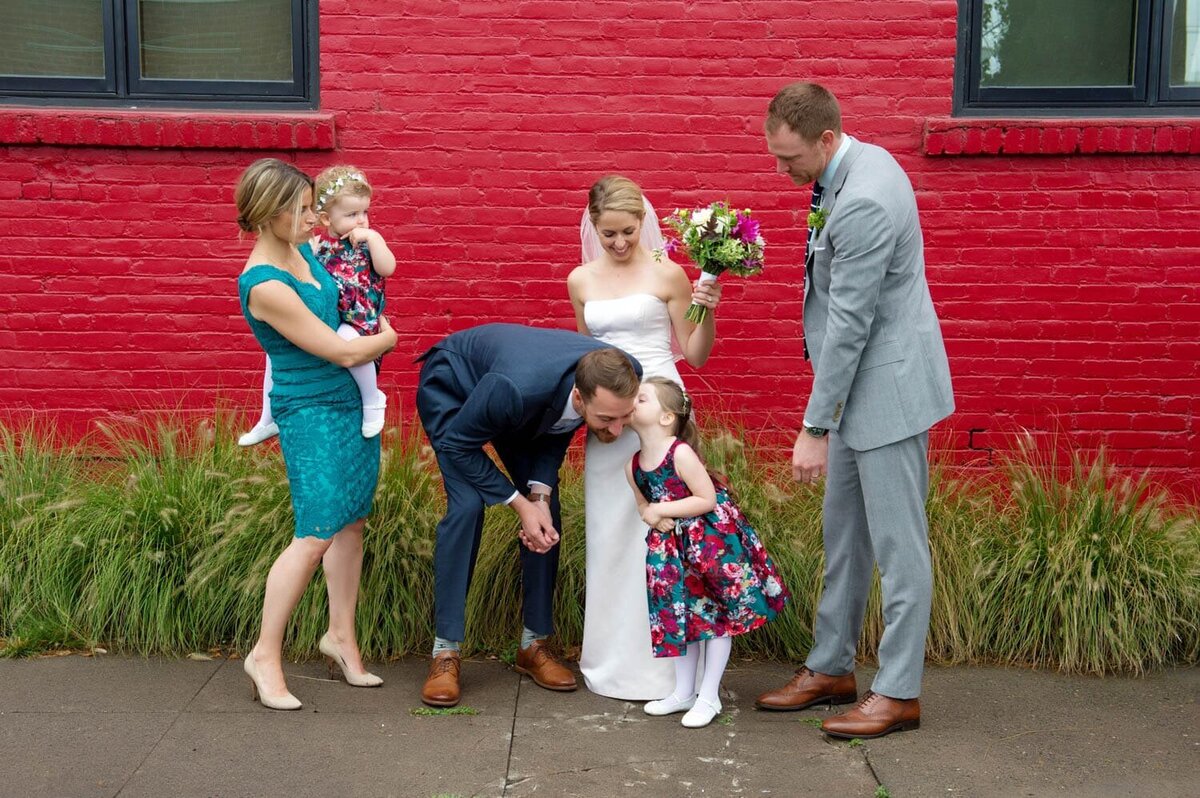 a groom leans down so a flower girl can kiss his cheek in front of a vibrant red wall