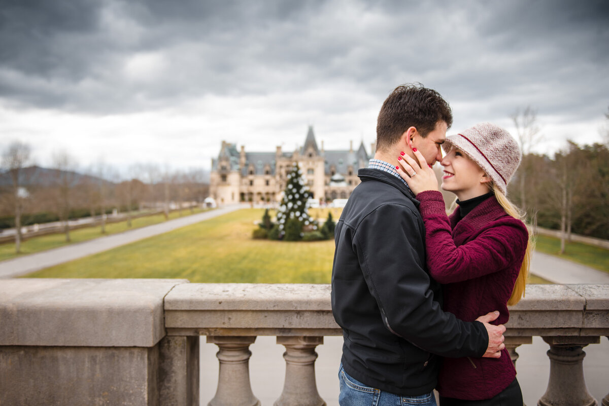 A-newly-engaged-couple-smiling-with-their-foreheads-together-her-new-ring-from-the-proposal-that-just-took-place-showcased-with-the-Biltmore-Estate-in-the-background
