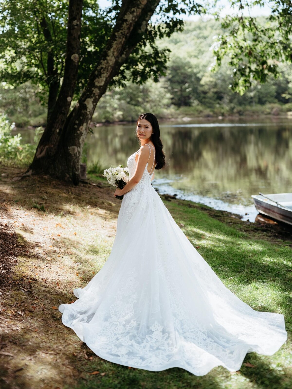 Bride poses for elegant portrait showing her dress, train, and bouquet under a tree, next to a rowboat in front of a lake and wooded area.