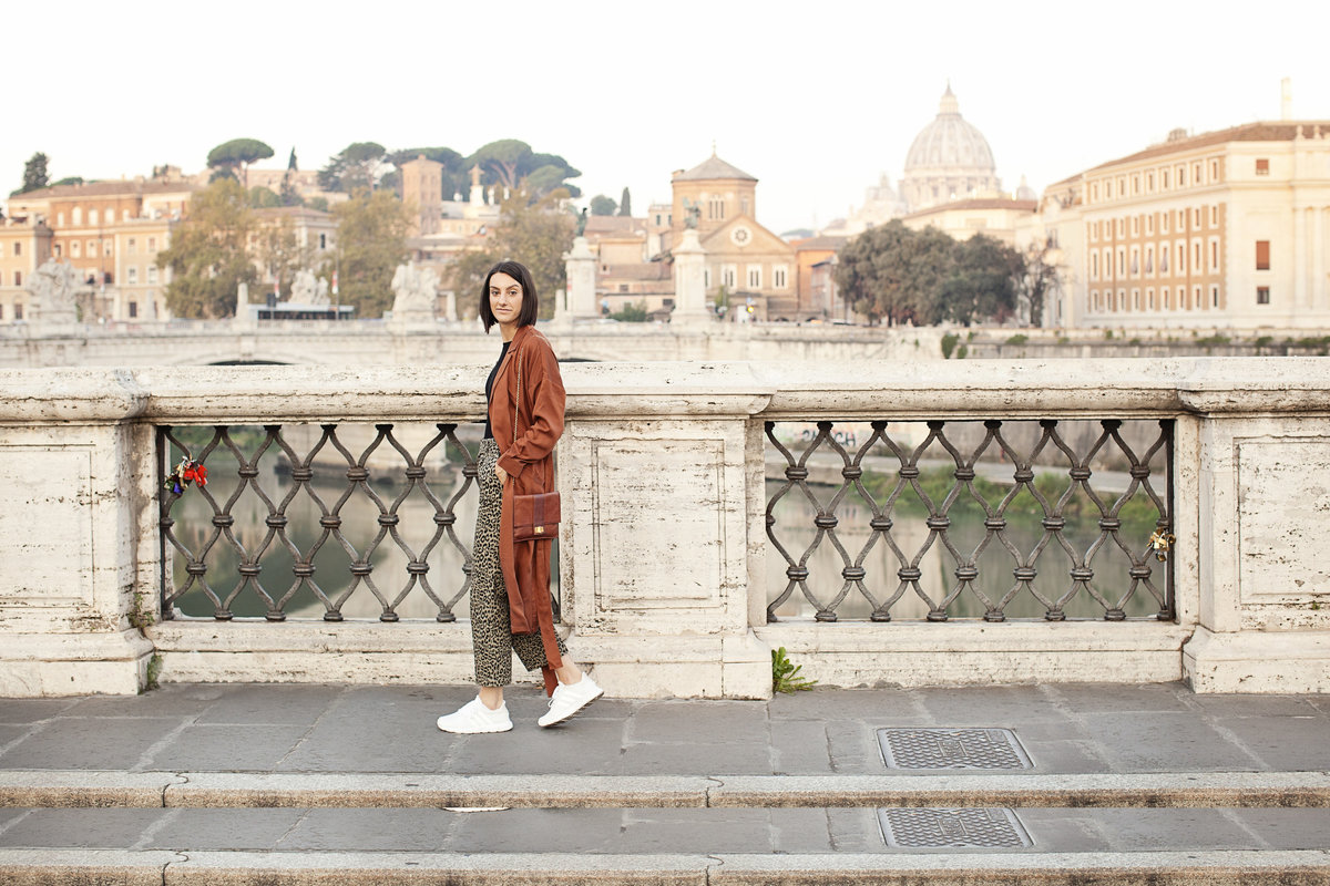 A stylish girl in an orange coat  walking on the Ponte Sant'Angelo bridge with views of Vatican City in the distance. Taken by Rome Photographer, Tricia Anne Photography.