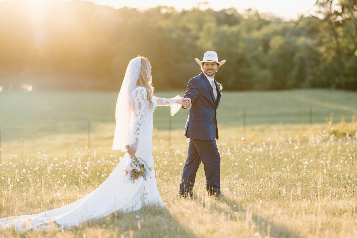 Sunset wedding portrait of East Tx bride in long traditional white wedding dress walking through open field with groom in navy suit and cowboy hat