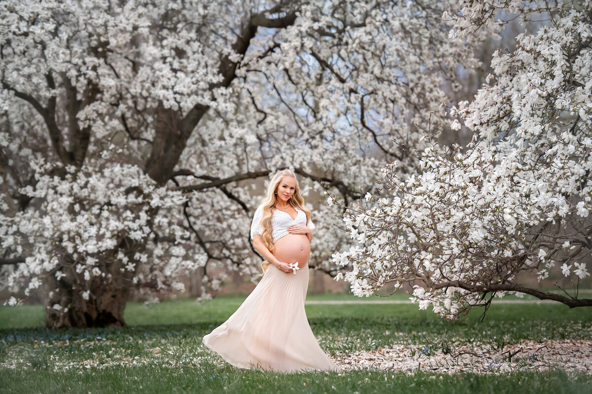 Barbie look a like with super long blond hair, surounded by spring magnolia blossoms, is holding her belly with baby number three.