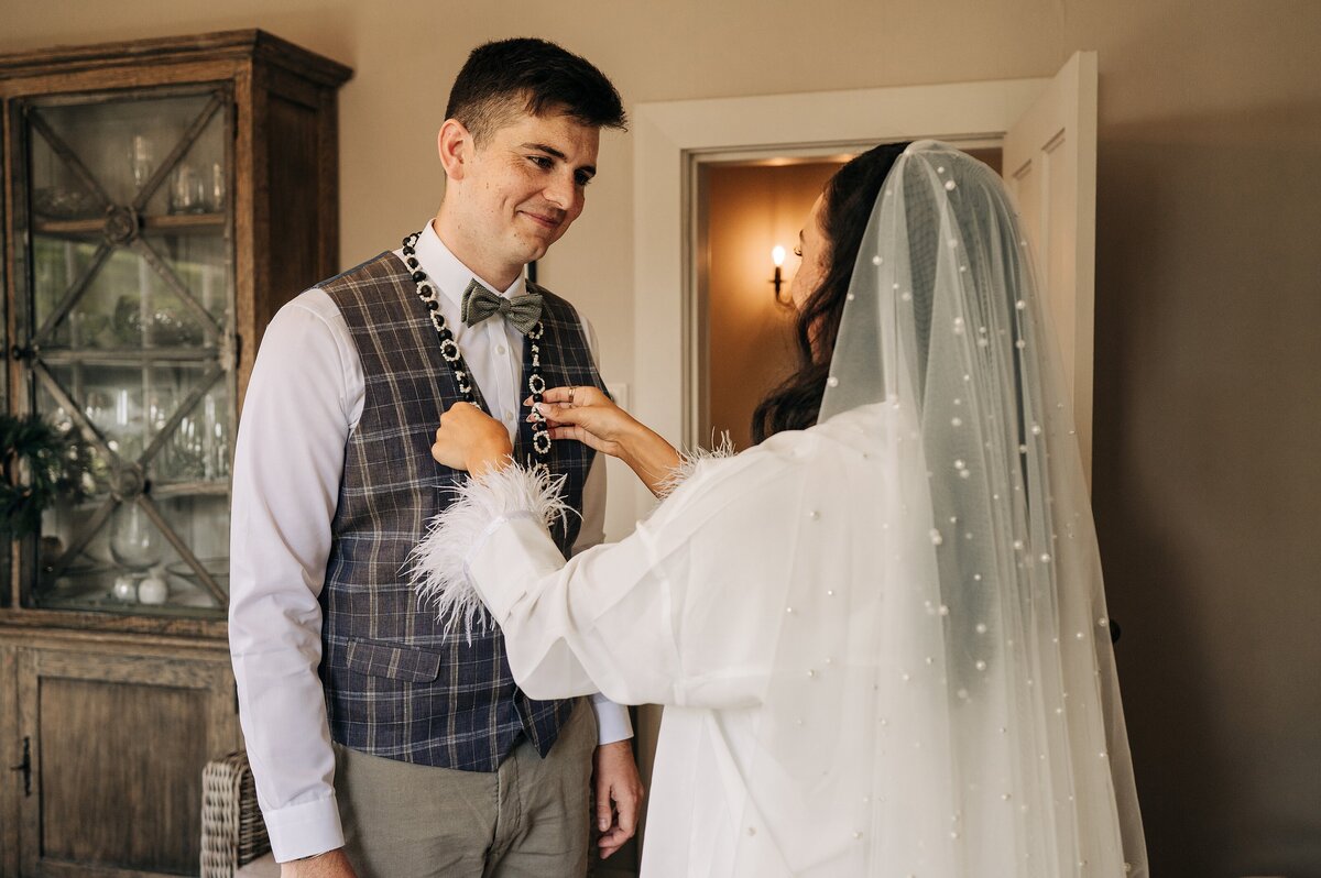 bride in white pearl veil puts ei pupu rarotongan beads on groom in checked waistcoat in christchurch house