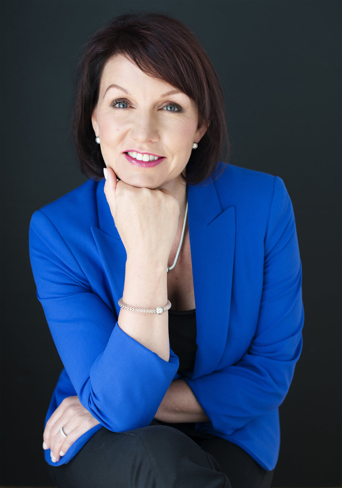confident headshot of  business woman sitting on chair with hand  under chin wearing blue jacket looking into camera  photograph taken by oakville best photography studio