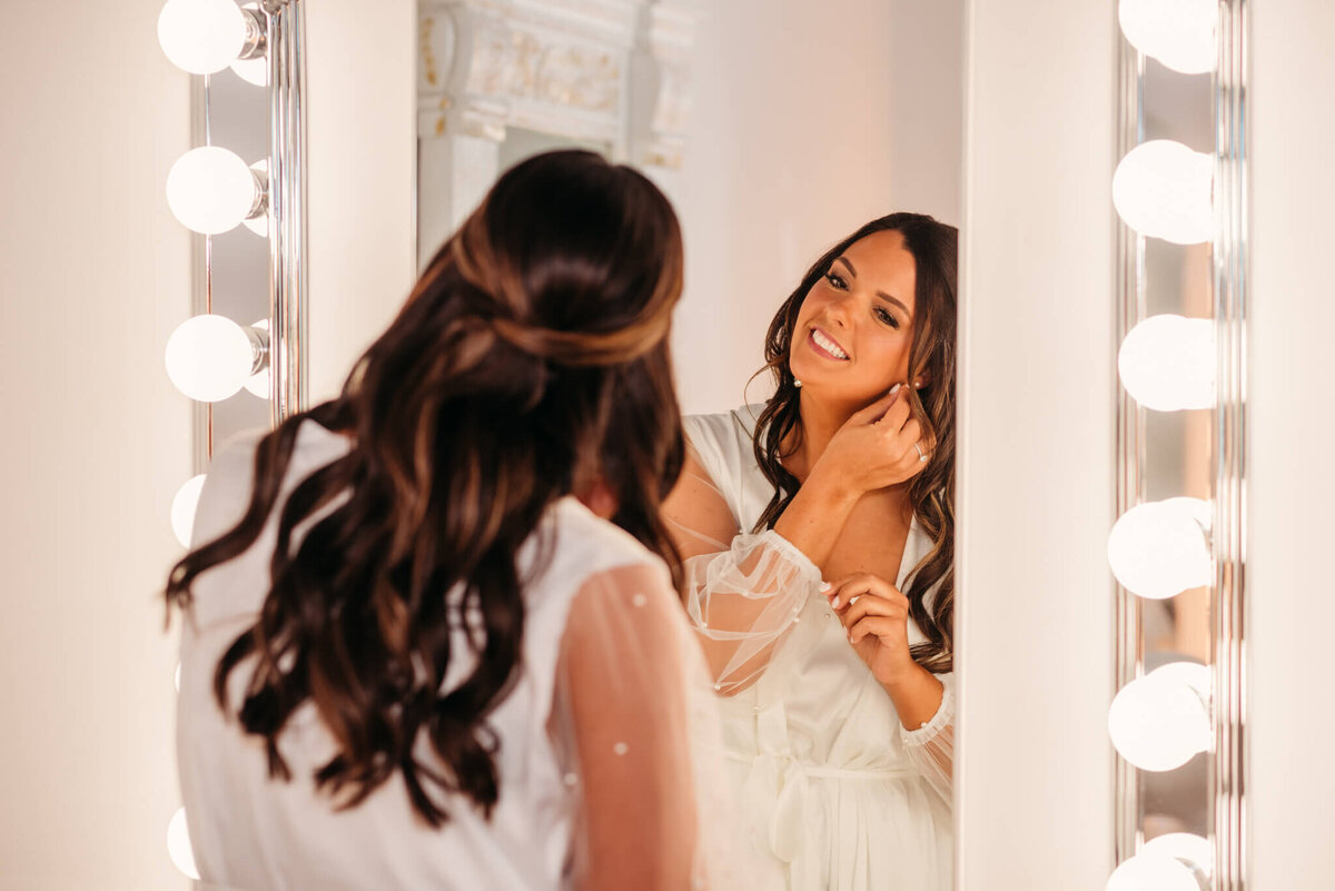 photo of a bride in a white robe standing in front of a mirror and putting in earrings