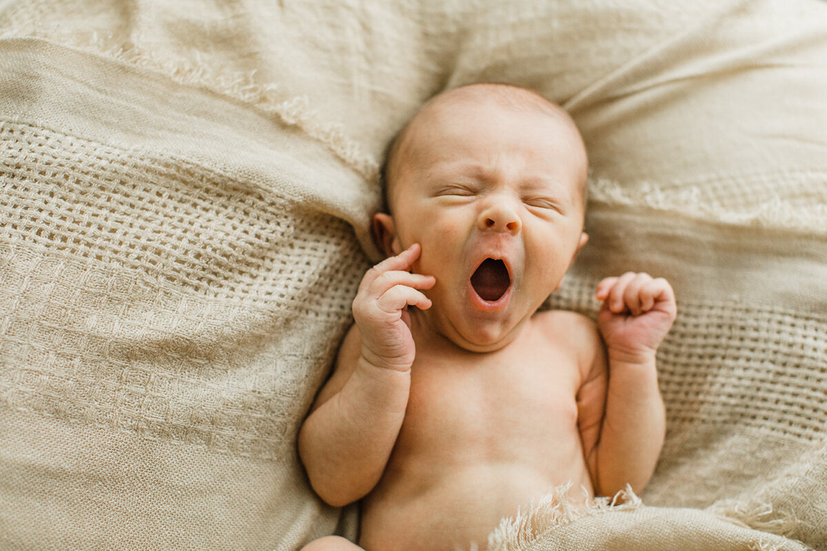 newborn baby lays on tan blanket and yawns