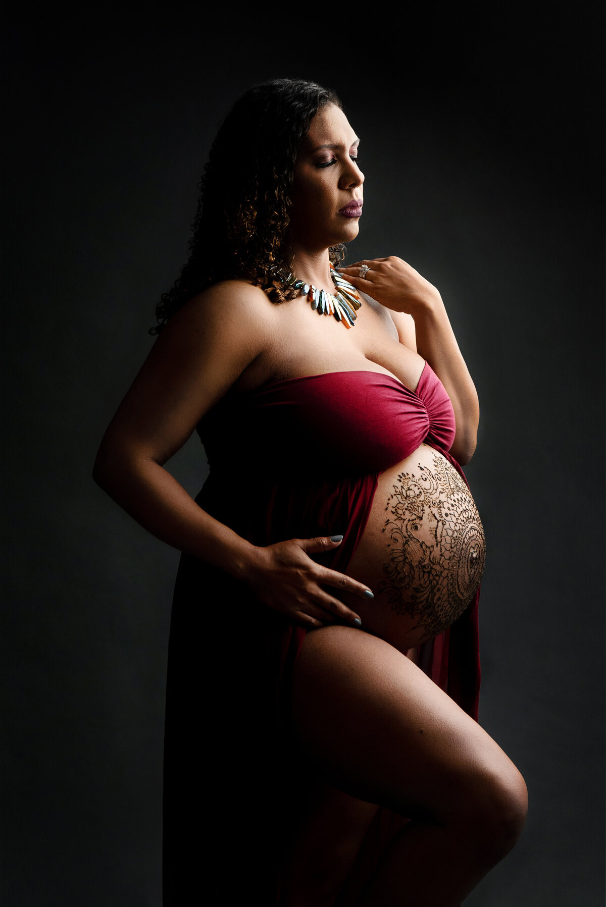 st-louis-maternity-photographer-expecting-mother-in-red-gown-showing-belly-with-henna-against-dark-background