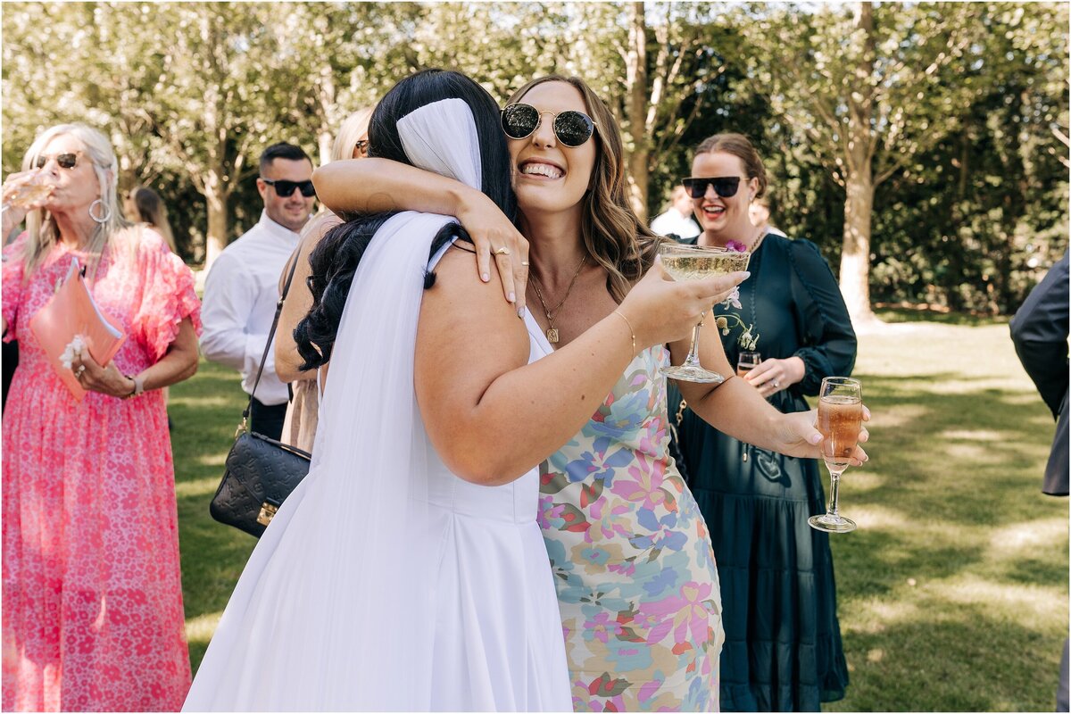bride hugging friend at wedding loving ellies belly stacey banfield holding champagne in shade of trees summer day