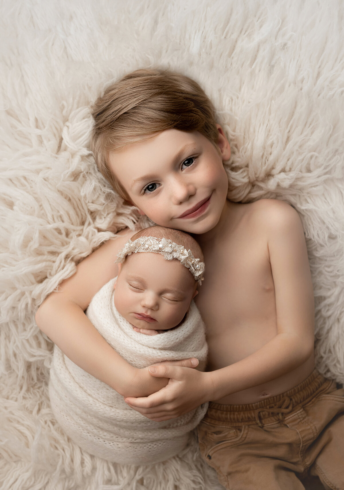 Philadelphia's best newborn photographer, Katie Marshall captures an aerial image of a bare-chested big brother holding his new baby sister. The brother is looking at the camera with his arms wrapped around baby. Baby sister is swaddled in cream with a matching lace and pear-embellished headband.