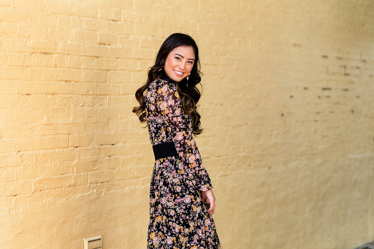 Richmond, VA senior girl looks over shoulder and smiles during her senior portrait session. Her session was in Uptown Richmond, VA in front of a yellow brick wall.