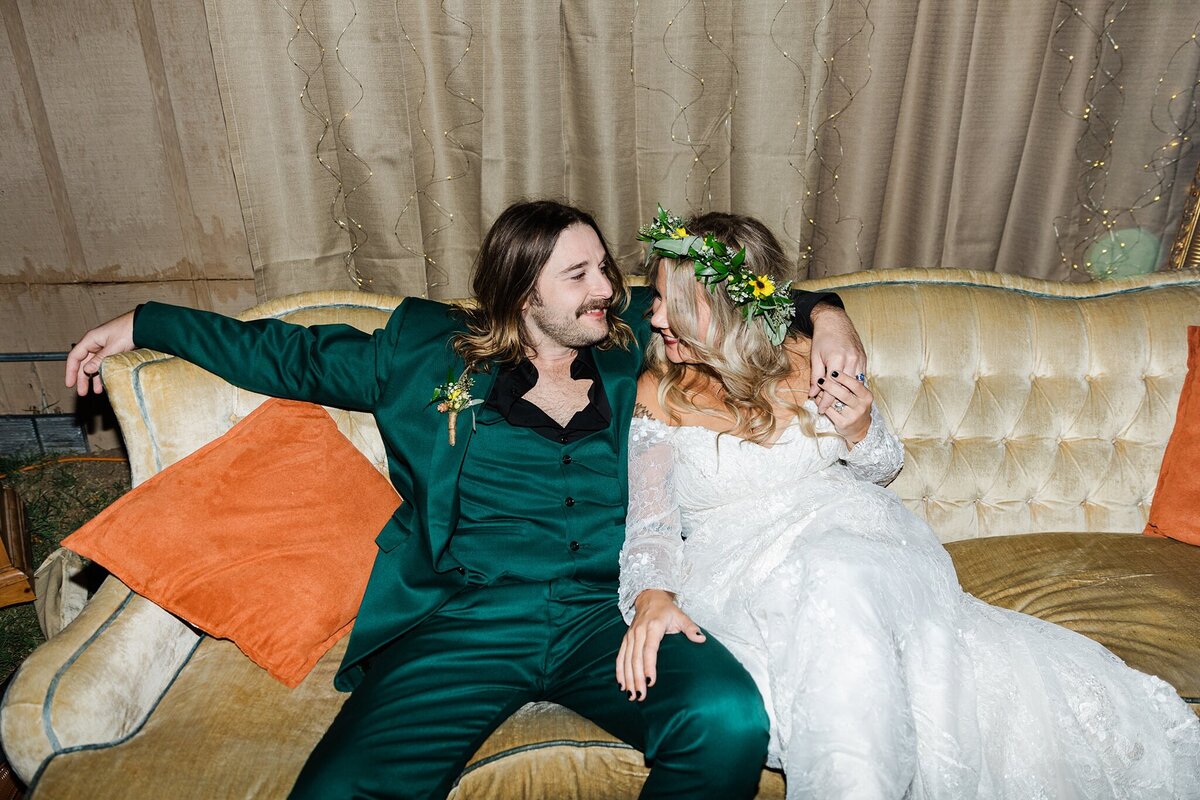 A bride and groom relaxing on a vintage couch during their outdoor, backyard, wedding reception in Fort Worth, Texas. The bride is on the right and is wearing a long sleeve, intricate, white dress and a flower crown. The groom is on the left and is wearing a green suit with a boutonniere.