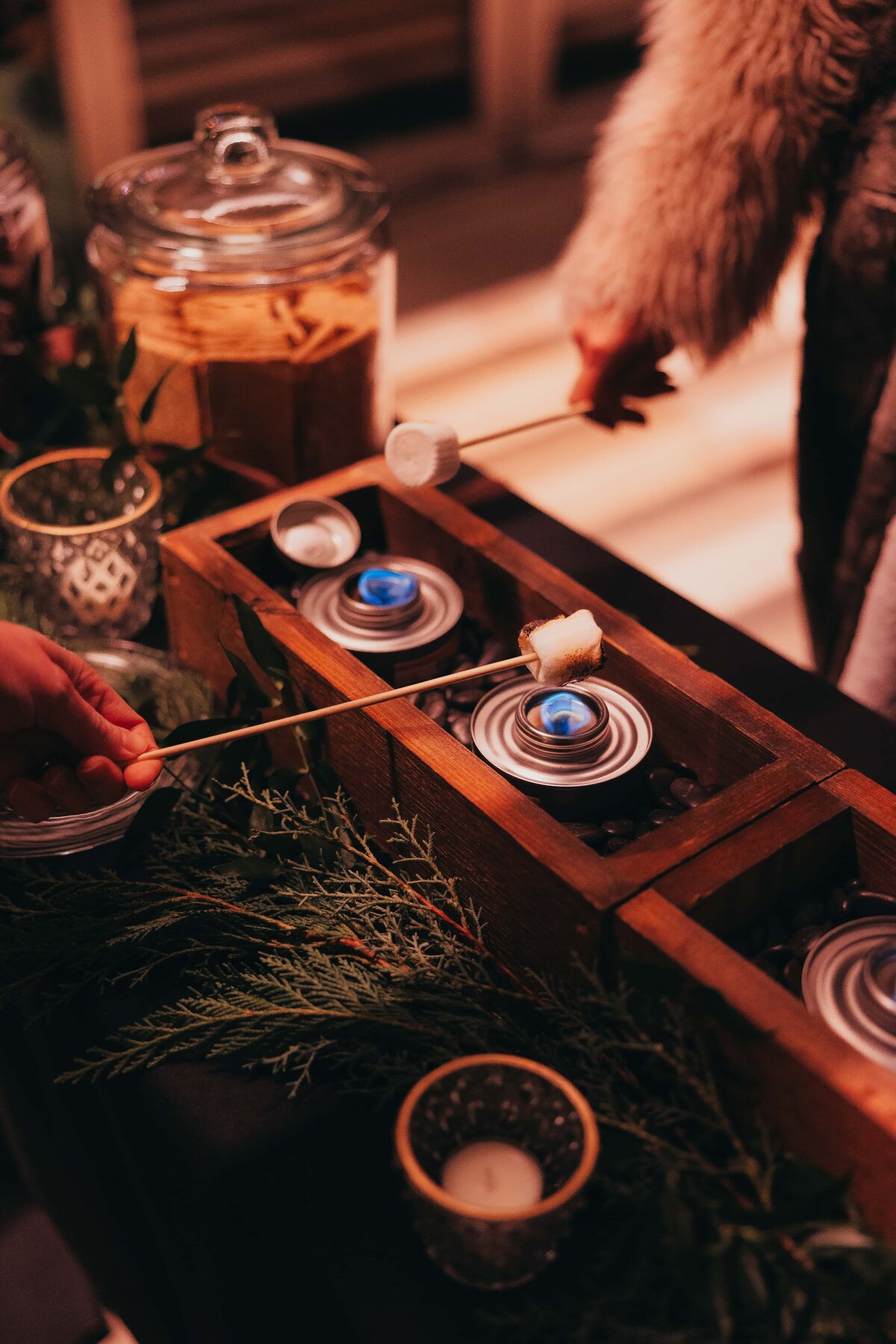 A person toasts a marshmallow over small candles at a cozy outdoor serving station adorned with greenery during an Iowa wedding.