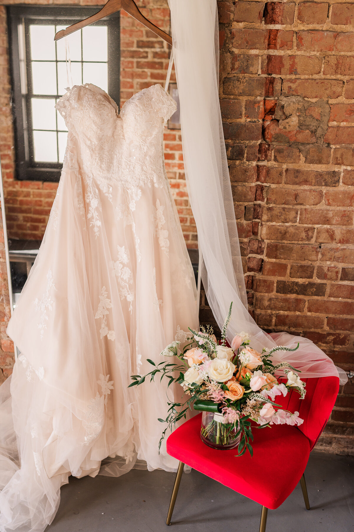 A cream wedding dress hanging on a mirror in a natural brick bridal suite by JoLynn Photography
