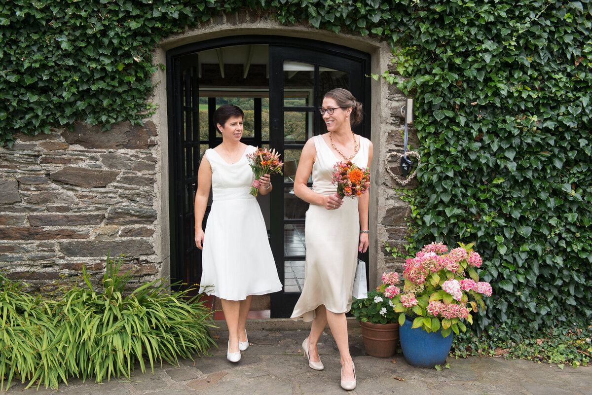 two brides wearing white dresses, walking and laughing while holding orange bouquets