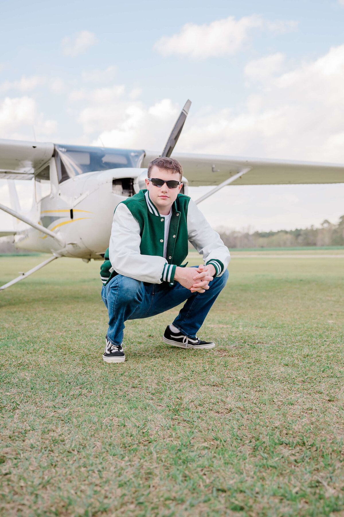 A person squatting on grass in front of a small airplane. They are wearing sunglasses, a green and white varsity jacket, blue jeans, and black sneakers. The sky is partly cloudy.