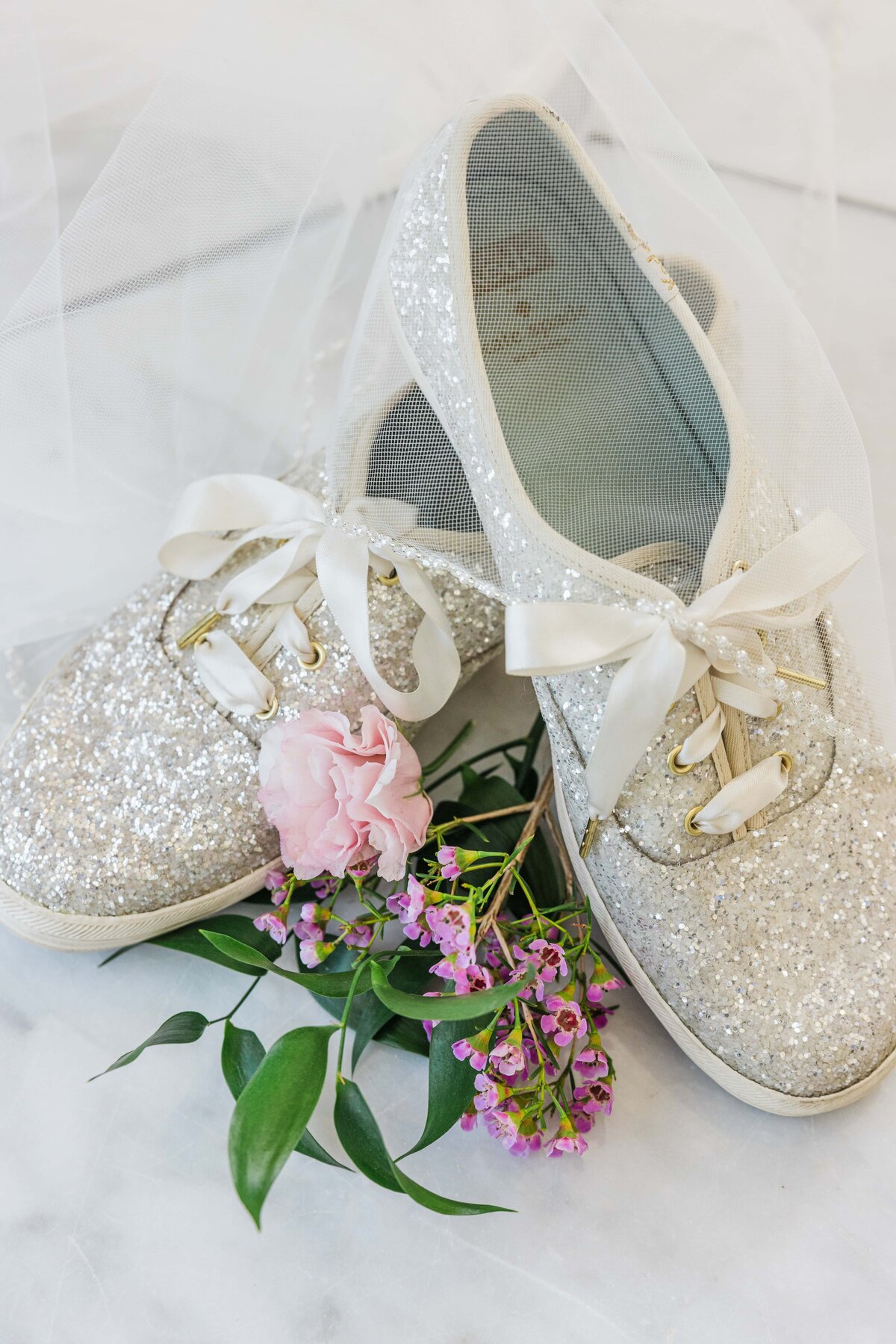 A pair of glittery bridal shoes adorned with bows, accompanied by a small bouquet of pink and purple flowers on a marble surface, perfectly curated for a Park Farm Winery wedding.