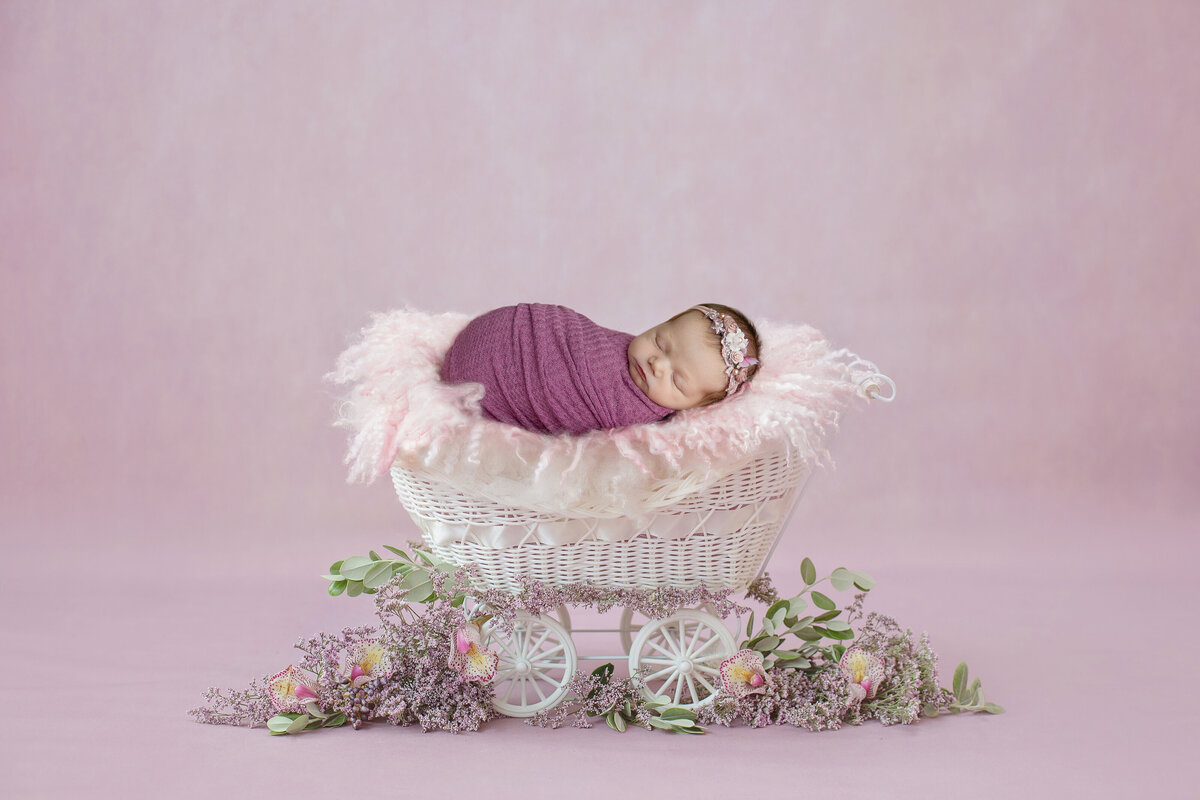 A newborn baby sleeps in a pink swaddle in a white wicker carriage in a studio