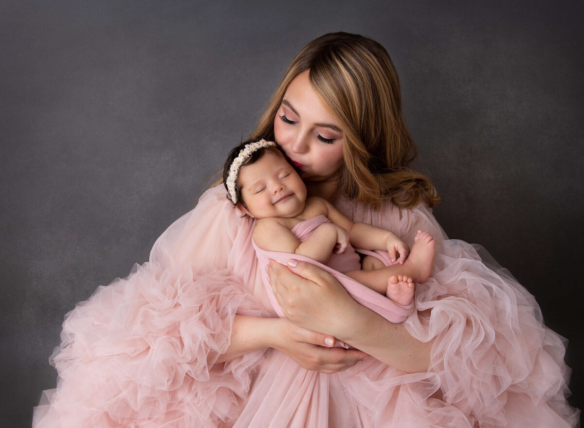 Mom in blush tulle gown is posing with her new baby girl, swaddled in a pink wrap with her hands and fingers exposed. Mom is kissing baby's temple and baby is smiling contently. Captured by Rochel Konik, top Brooklyn, NY newborn baby photographer.
