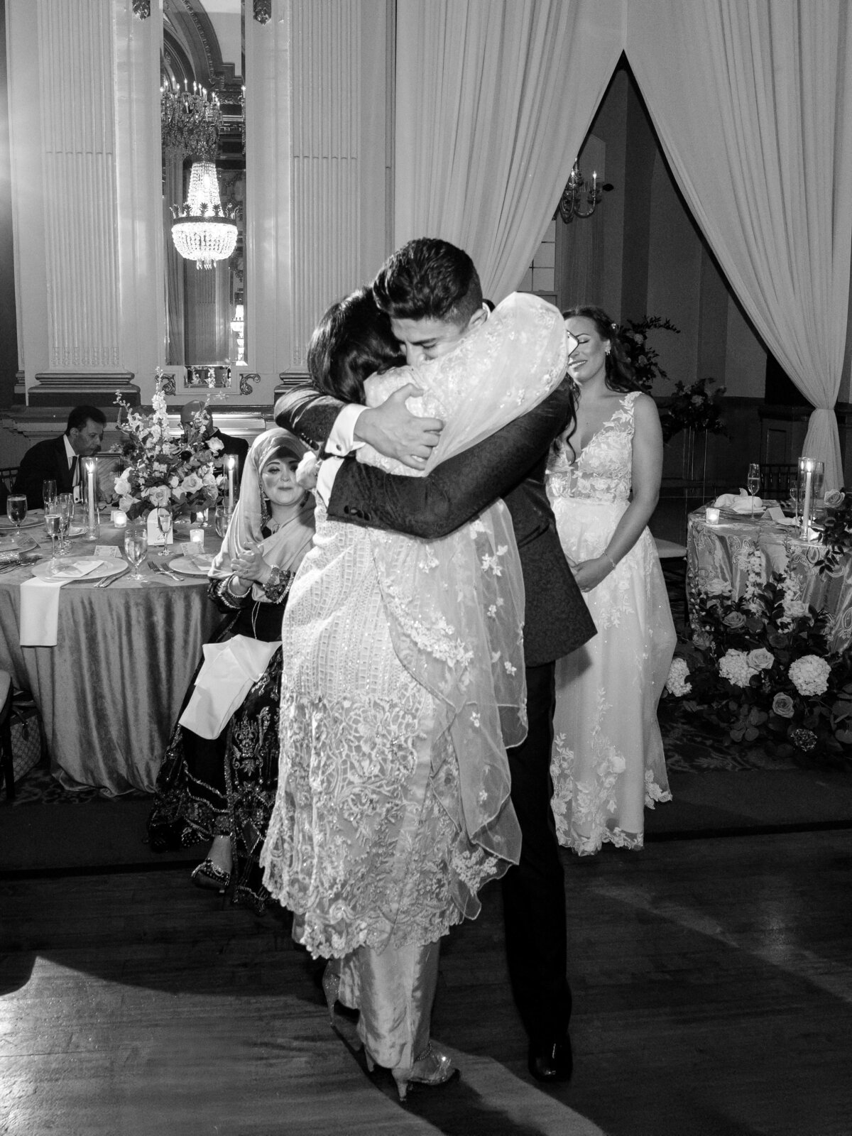 A black and white photo of a wedding reception. A groom warmly hugs a woman while a bride stands nearby, smiling. Guests sit at a table adorned with flowers in the background, and a draped archway is visible.