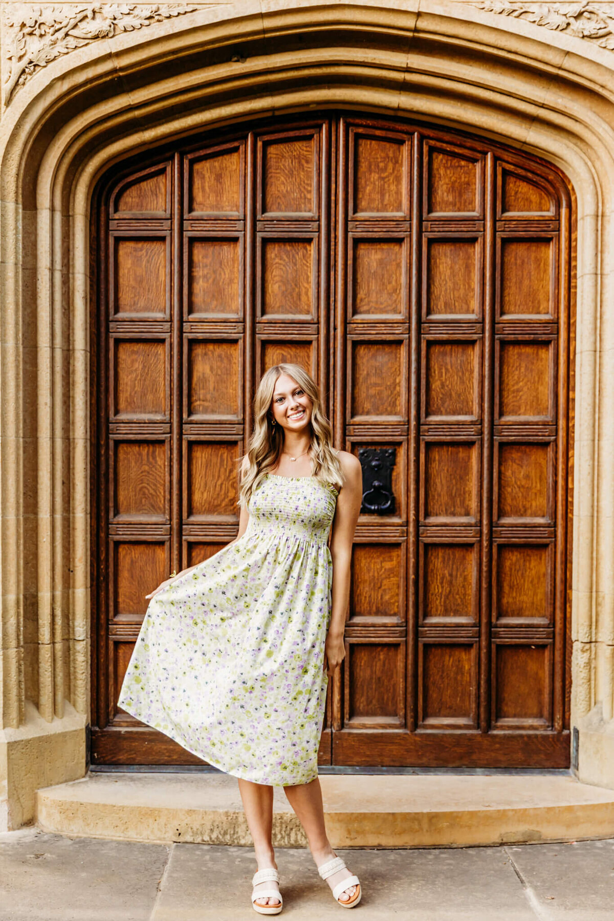 photogenic teenage girl playing with her floral pattern dress in front of gorgeous arched wooden doors for her senior photos