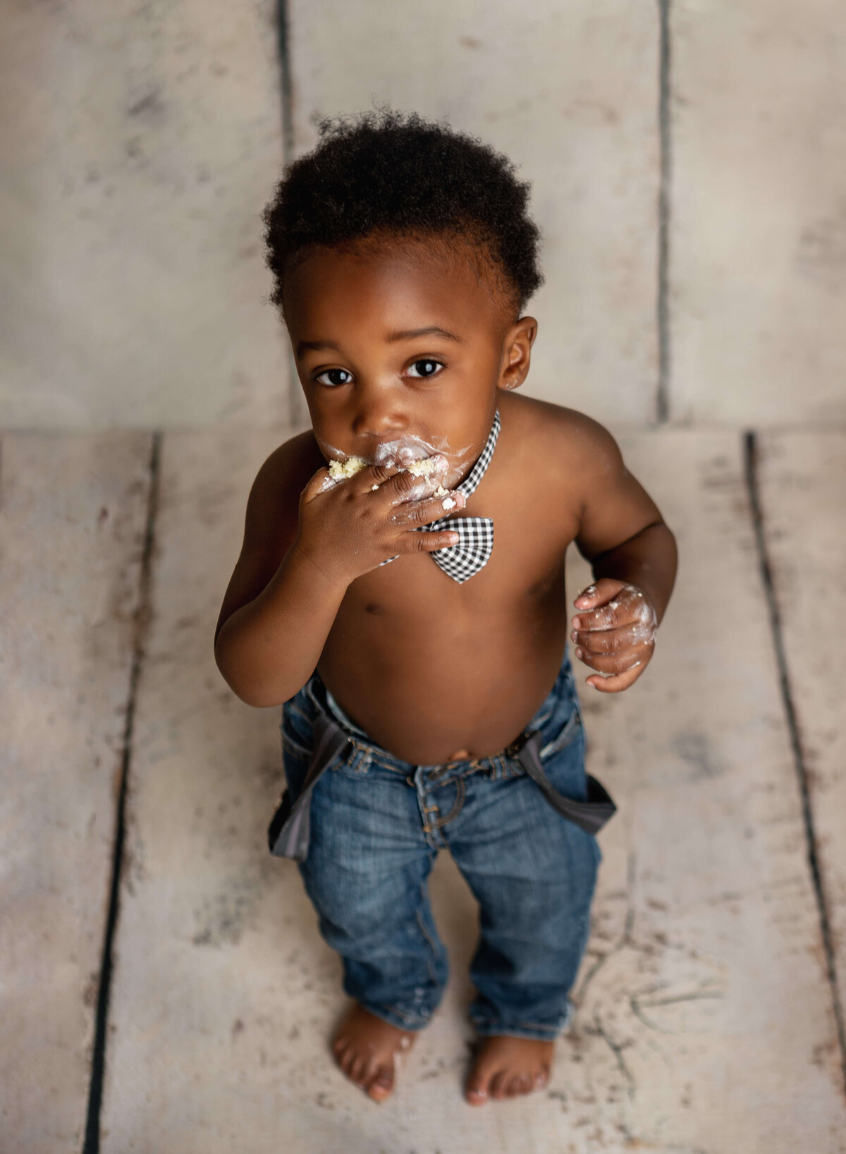 little boy standing eating cake in jeans and a bow tie on a white wood background in a cake smash session