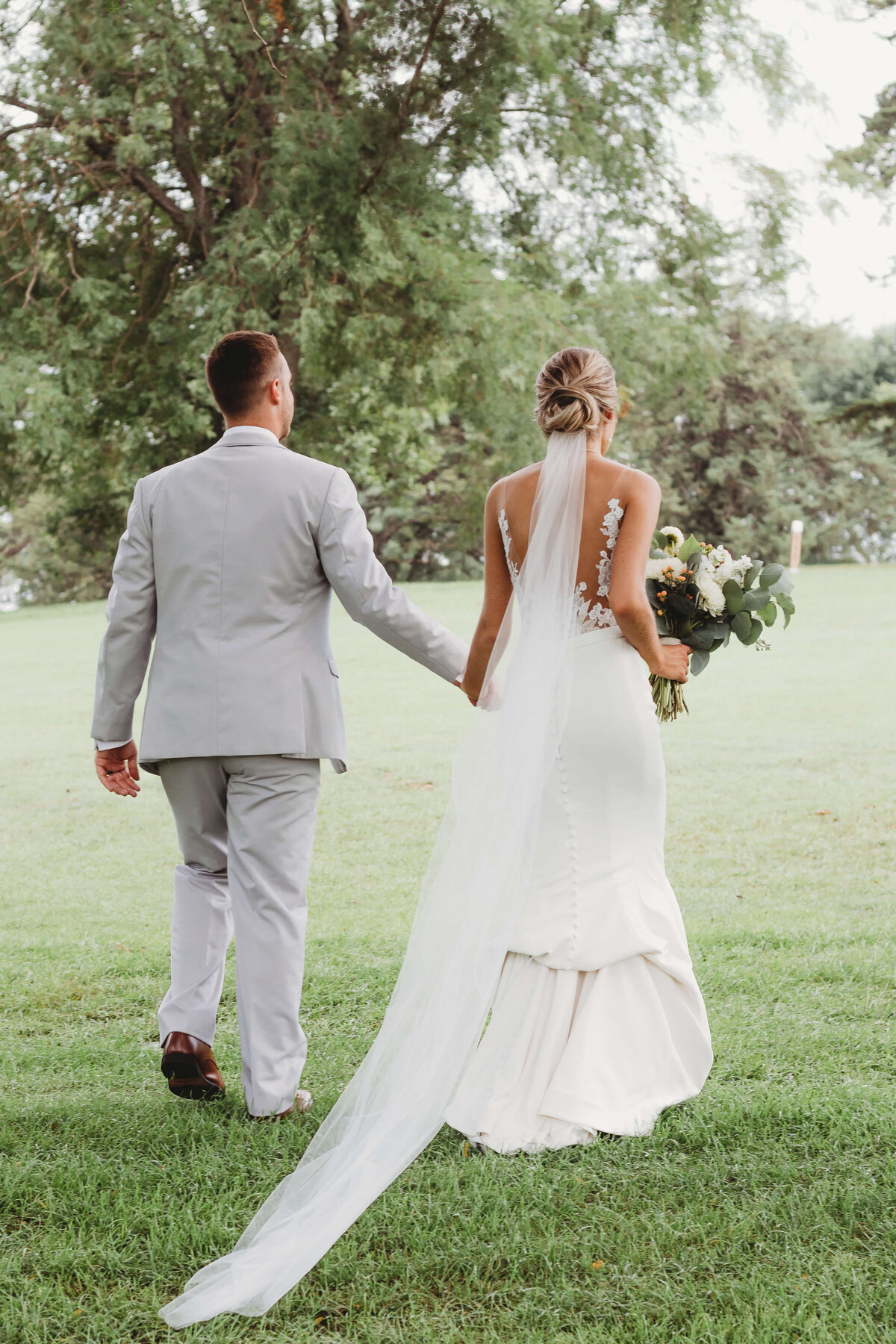 Husband and wife holding hands and walking away on golf course with the brides long veil  dragging.