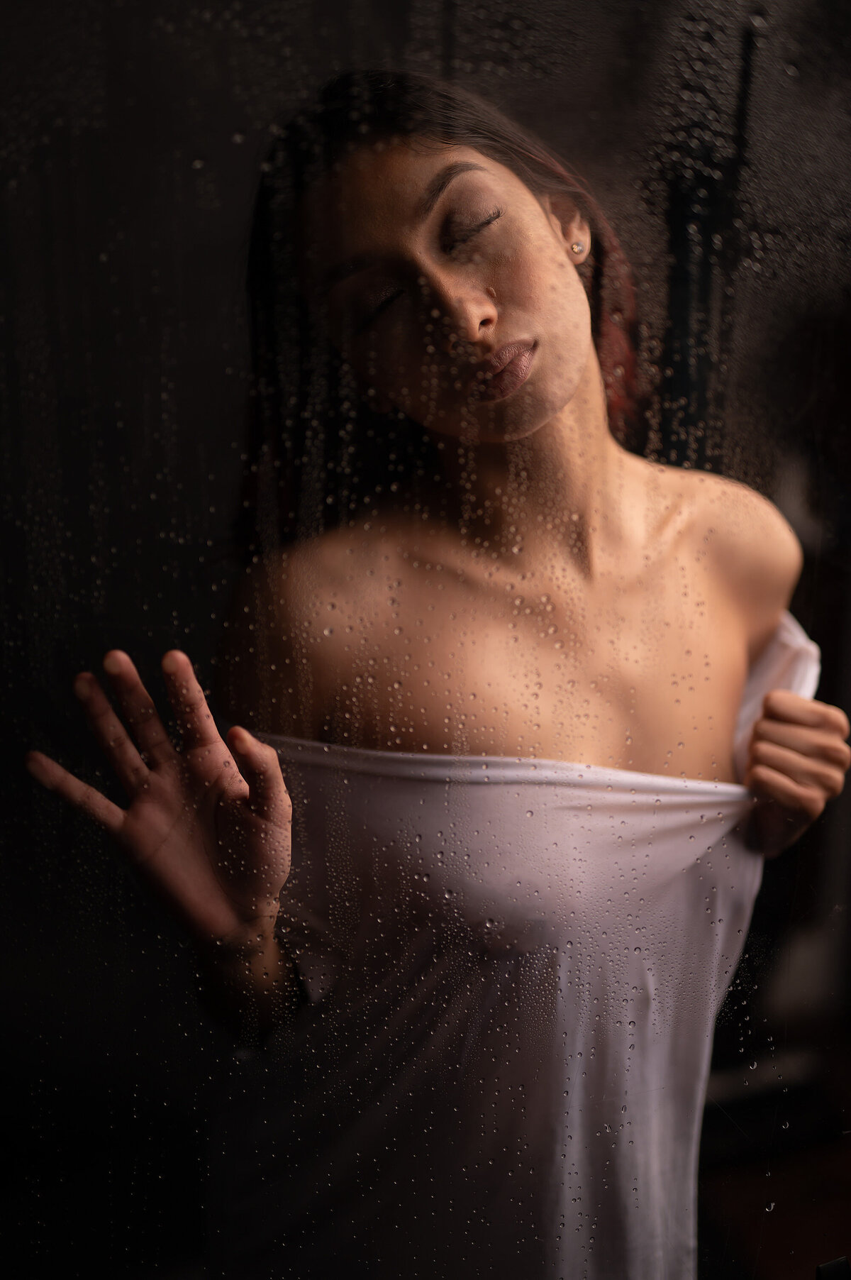 A sensual boudoir image of a woman in a sheer white top  behind a steamed glass panel.