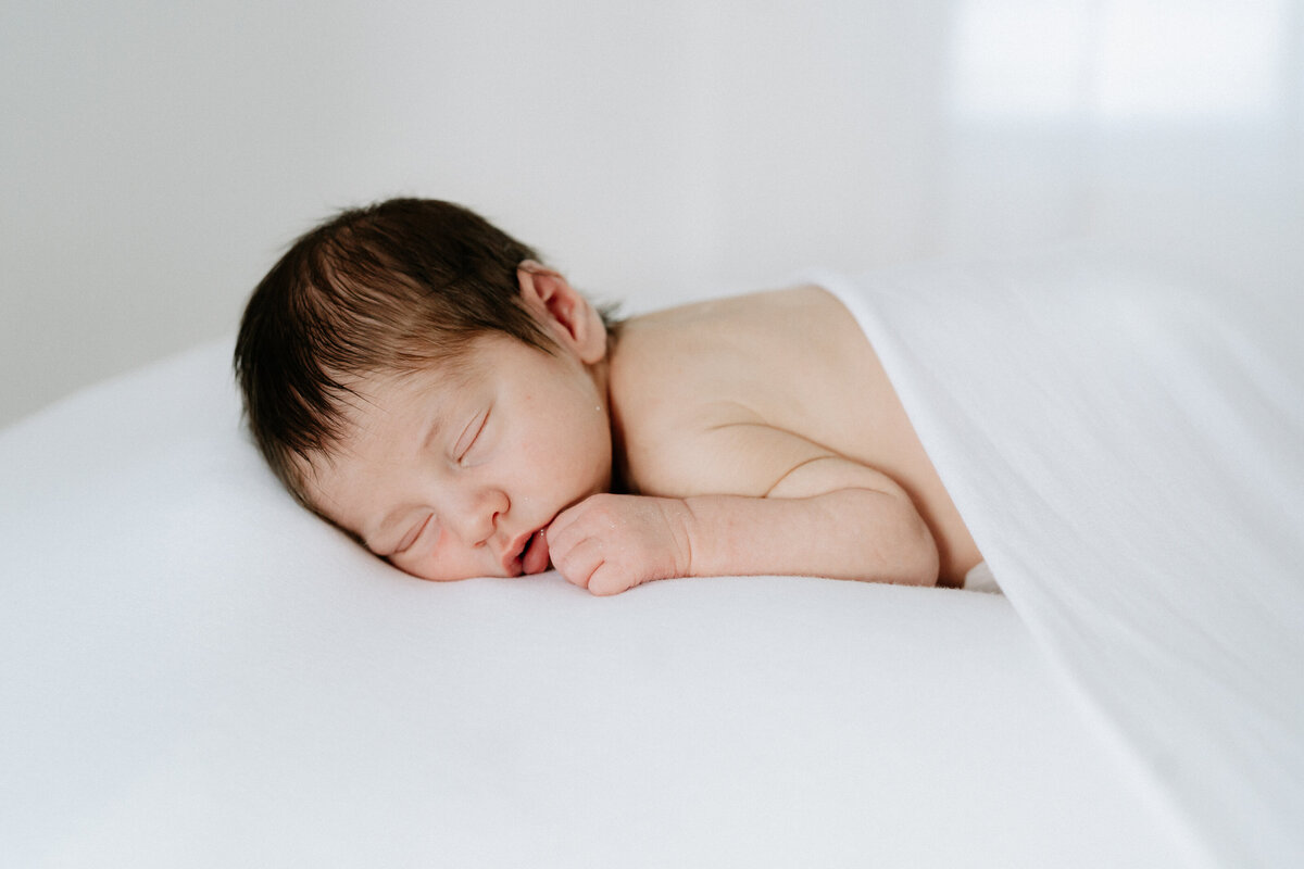 newborn baby boy wth brown hair sleeps under a white blanket during a newborn photo shoot