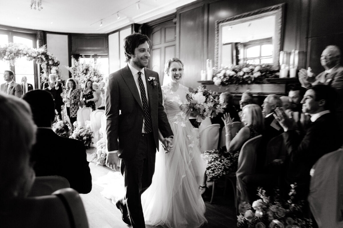 Black and white photo of the bride and the groom smiling while walking down the aisle as the seated guests clapped.