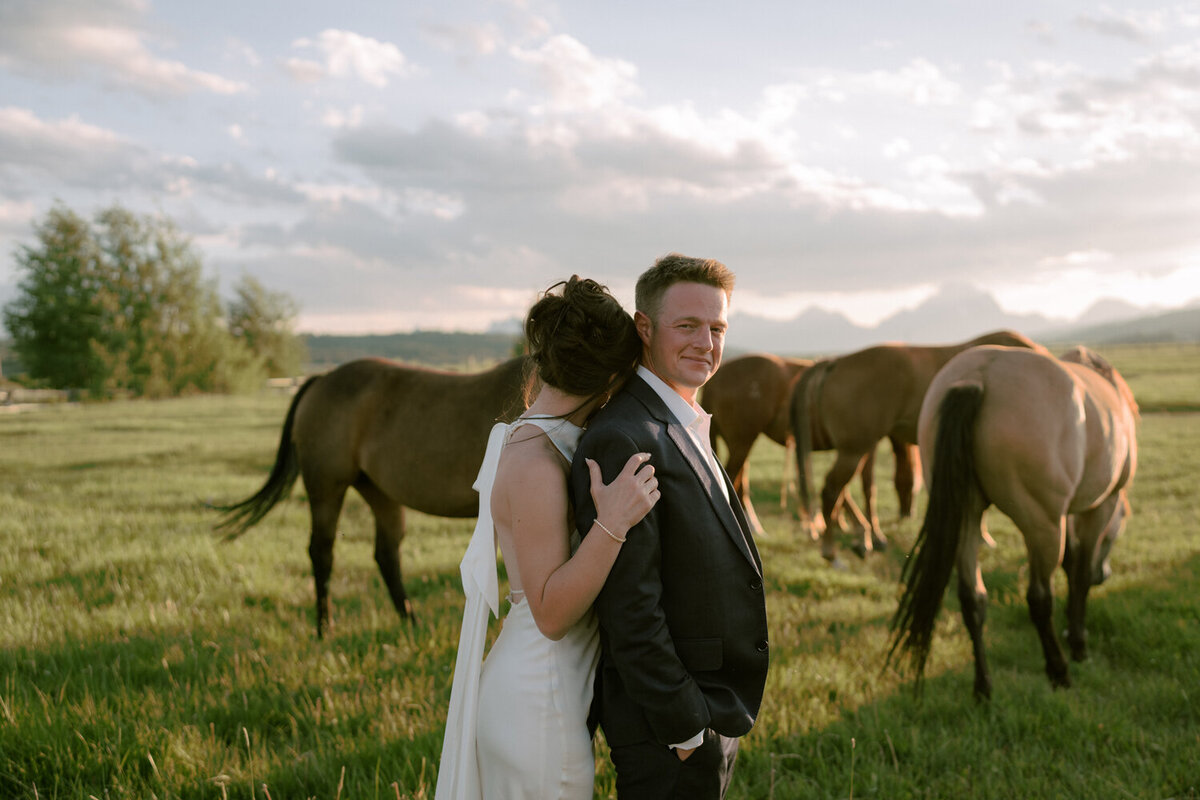Groom gazes confidently at the camera as his bride embraces him from behind, set against a summer backdrop of horses grazing in the rolling fields of Jackson, Wyoming.