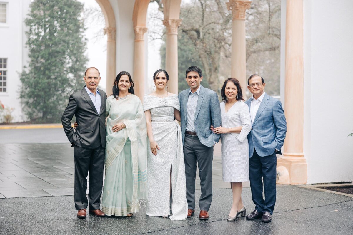 Three couples posing together in formal attire outside a building with columns.