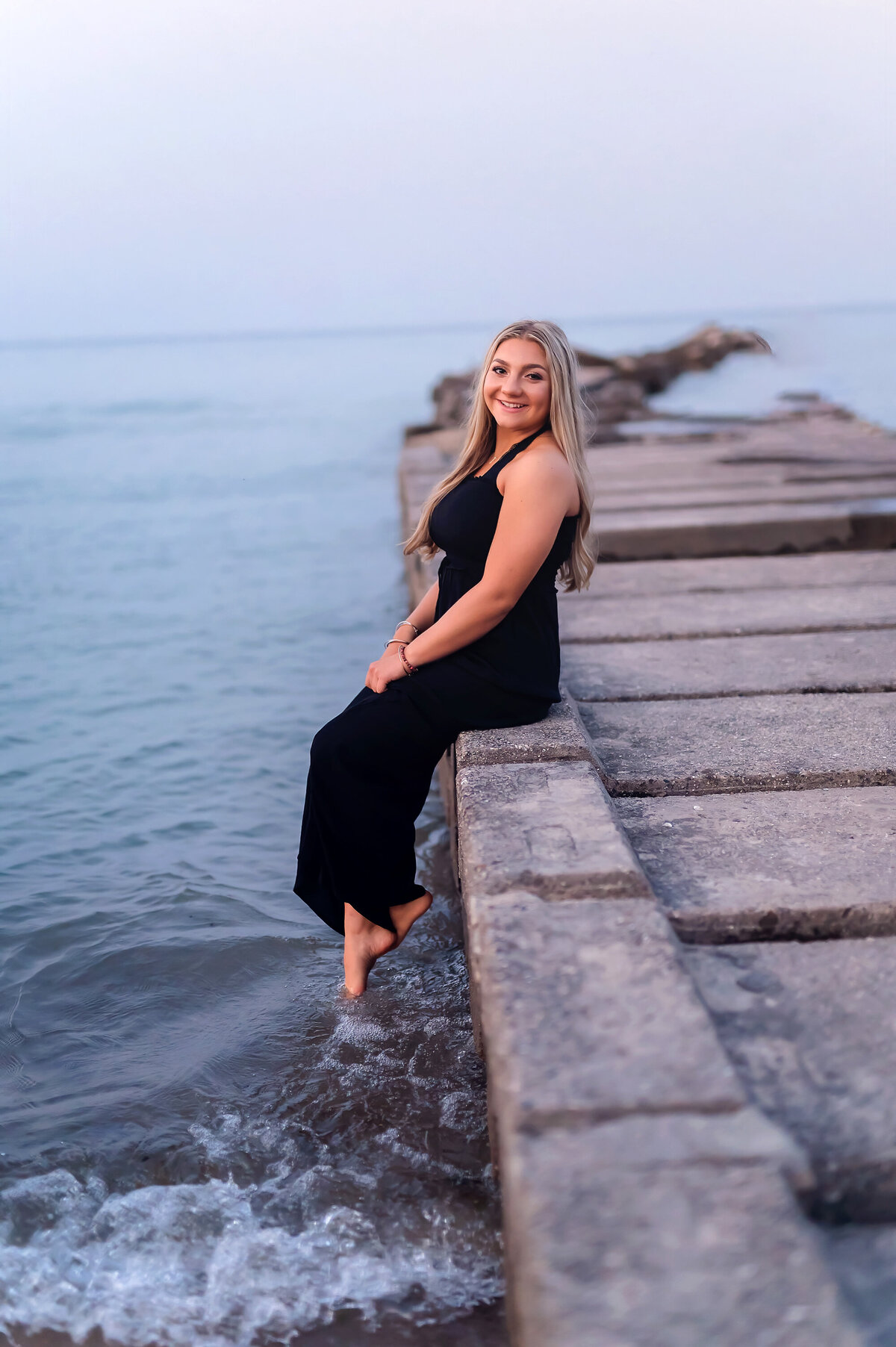 A female senior from Catholic Memorial High School sits on a breaker wall along Lake Michigan wearing a black jumpsuit for her senior photoshoot.