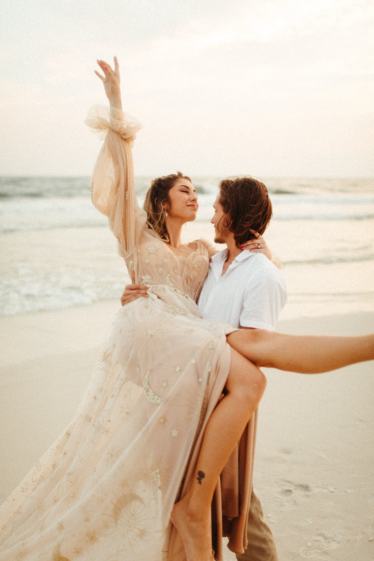 Groom holding bride on the beach as she throws her arm in the air and smiles after their elopement