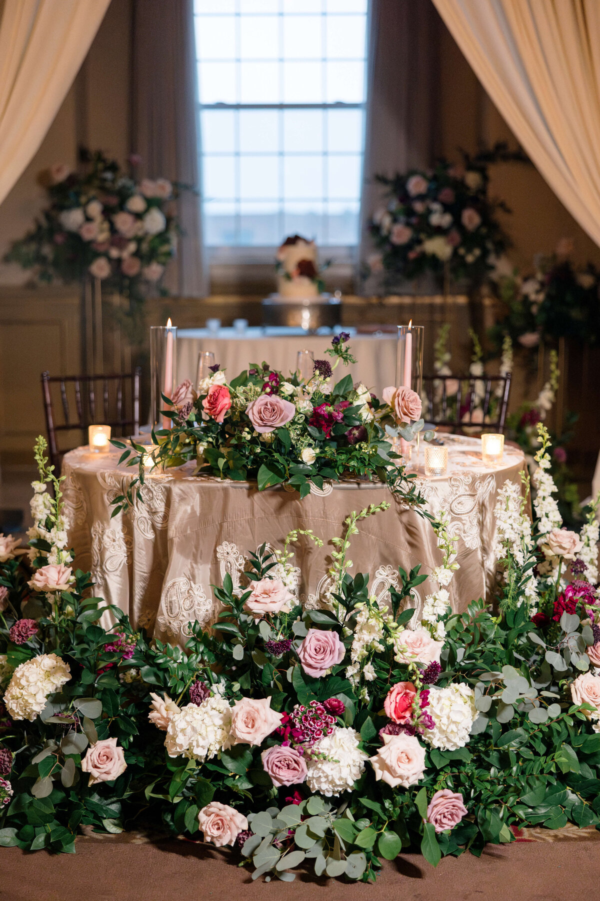 A beautifully decorated table for a wedding reception, adorned with elegant floral arrangements featuring pink and white roses, greenery, and candles. A large window and draped curtains provide a soft, romantic backdrop.