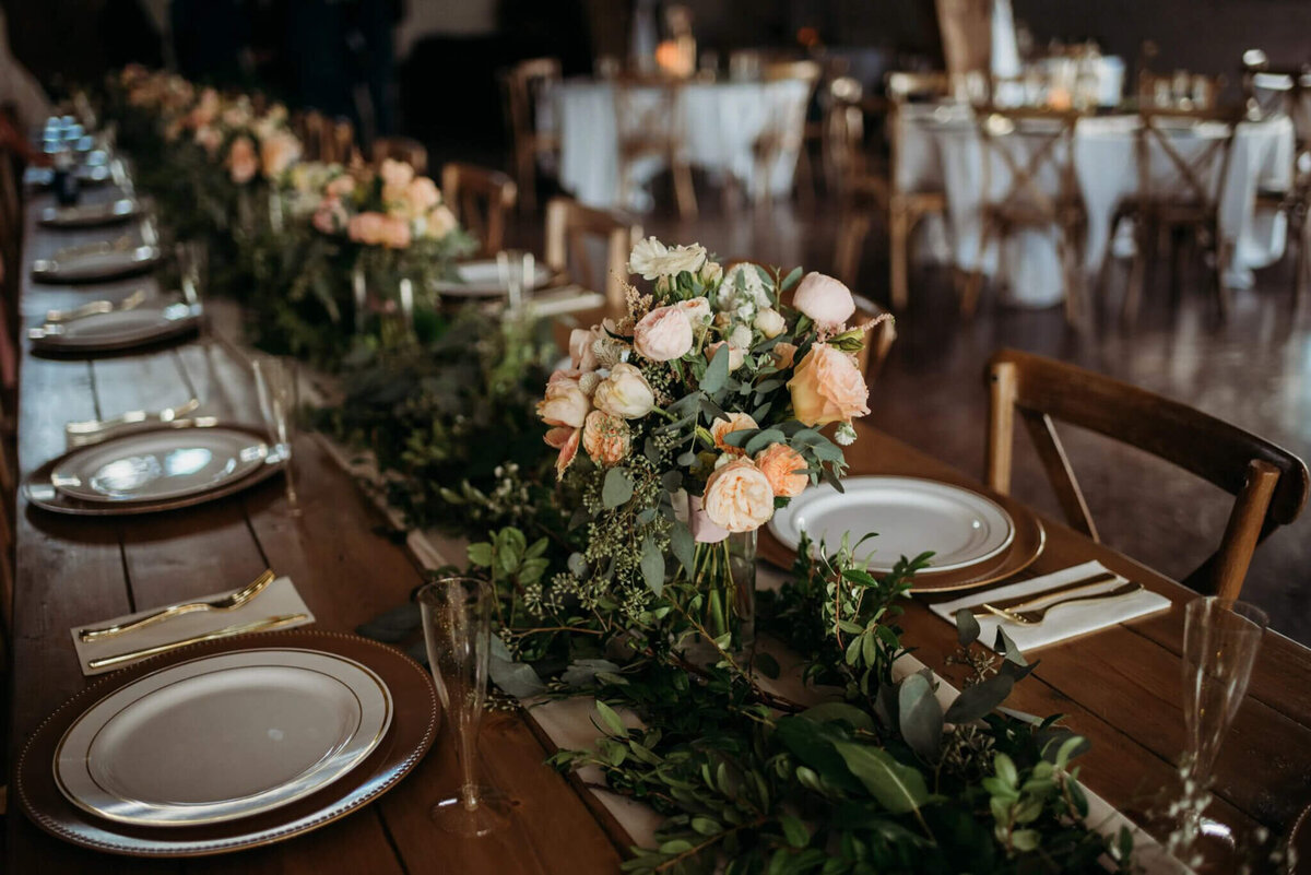 Photo of a wooden wedding table with greenery as the runner and pink flowers in vases