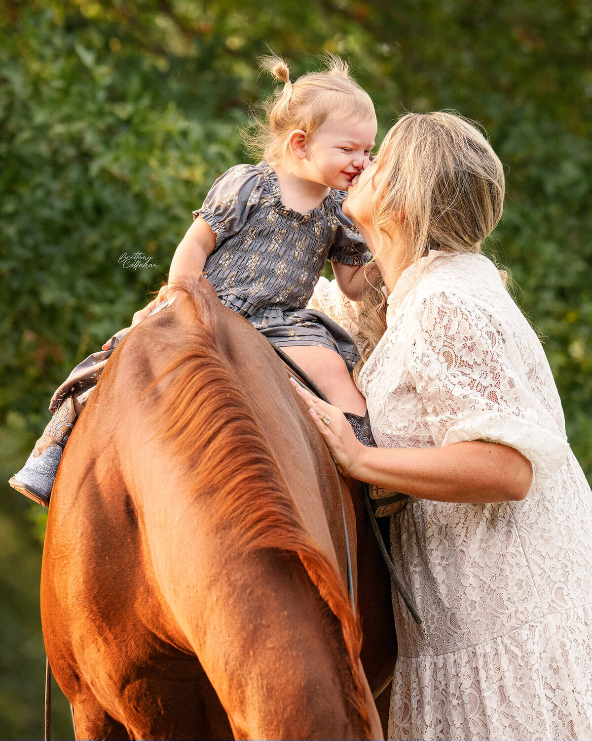 blonde equestrian in a lace cream dress photographed with her daughter sitting on their chestnut quarter horse giving each other a small kissl