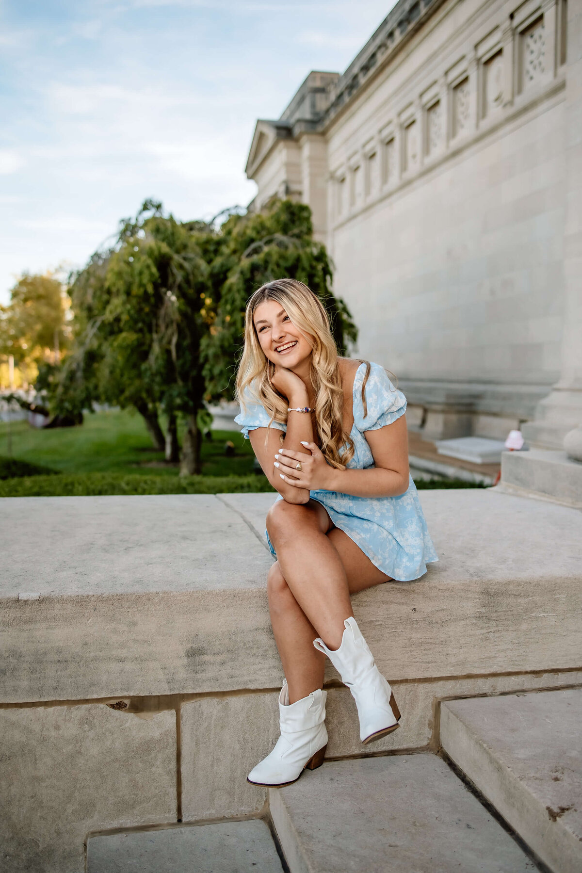 A blonde haired high school senior girl sitting and laughing on a ledge of a building