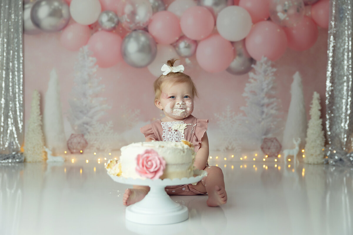 A happy girl covered in cake sits in a studio in a pink dress in a New Jersey Cake Smash Photographer studio