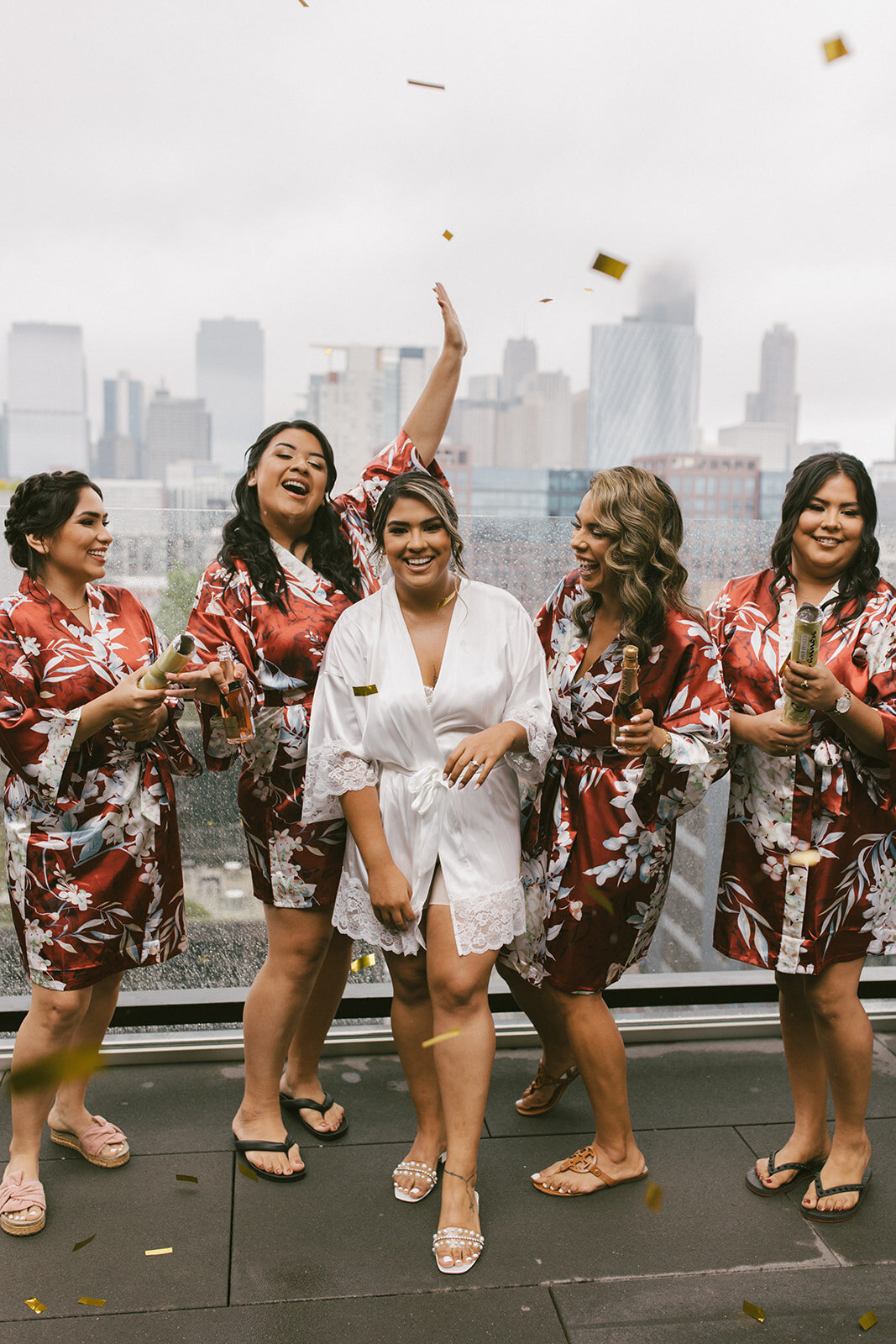Bride in white satin, lacy robe smiles at camera while four bridesmaids behind her in red and white floral robes shoot confetti overlooking Chicago skyline.