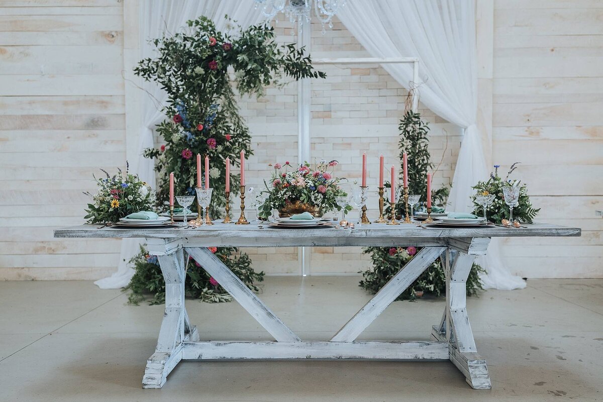 A white farm table against a white wood wall with white drapery is decorated with a lush asymmetrical  arbor and three small centerpieces of greenery accented with pink, purple, yellow and white wildflowers. The table is set with silver chargers and white plates that are topped with light blue napkins. Pink taper candles line the table in brass candle  holders.