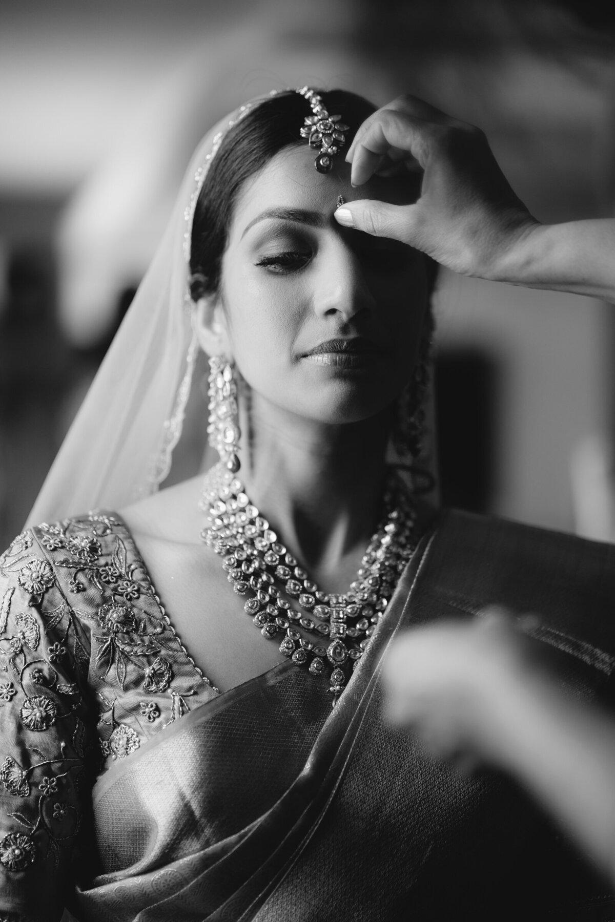 A black and white portrait of a South Asian bride, her expression serene as a hand adorns her with traditional bridal jewelry.