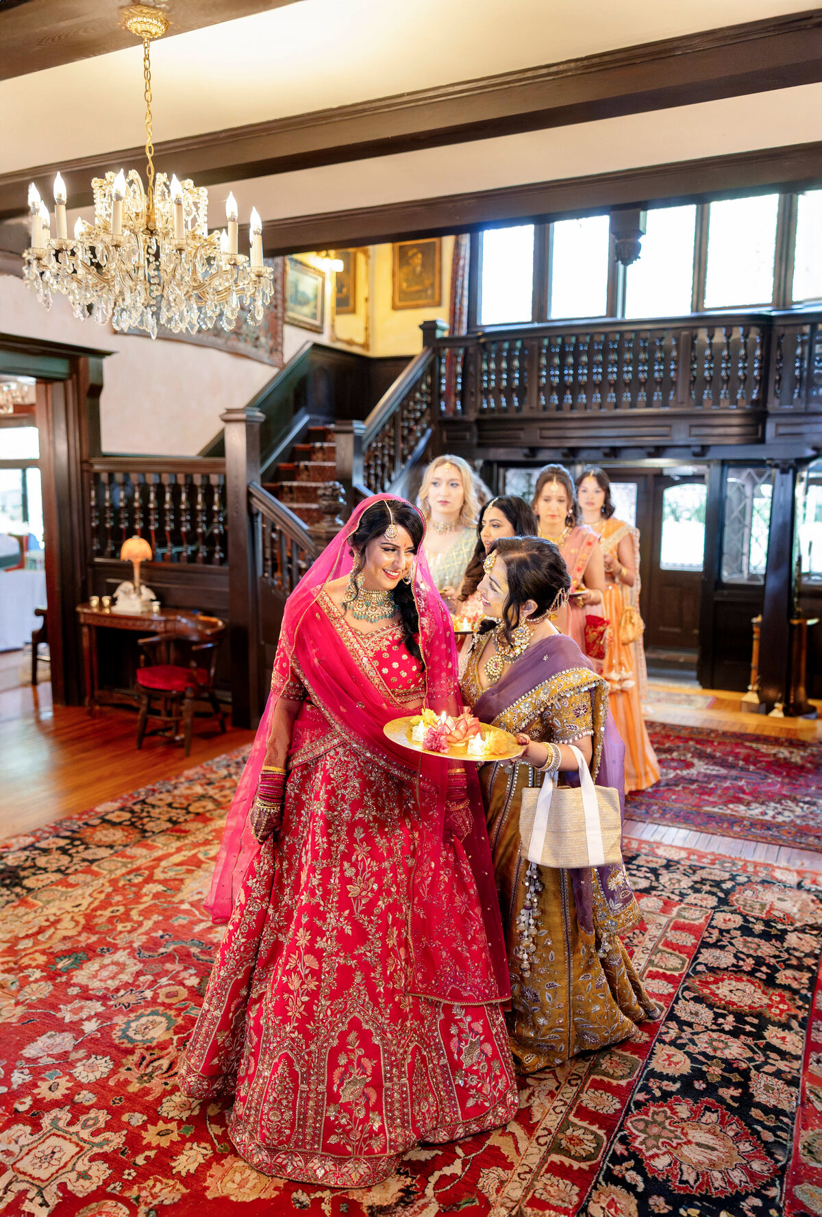 A bride in a vibrant red lehenga smiles as she walks with a procession of women in traditional attire, all holding a gold platter with offerings. They are in an elegantly decorated room with a chandelier and wooden staircase.