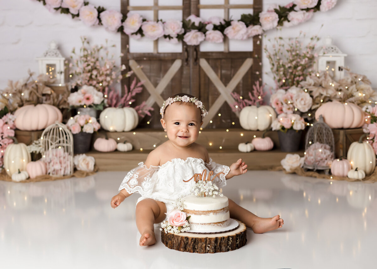 Elegant pink autumn themed cake smash in West Palm Beach and Boynton Beach photography studio. Baby girl is sitting in a boho off the shoulder dress smiling at the camera with a cake in front of her. She has not yet touched the cake and is very clean. In the background are shades of pink and cream pumpkins and a rustic barn door.