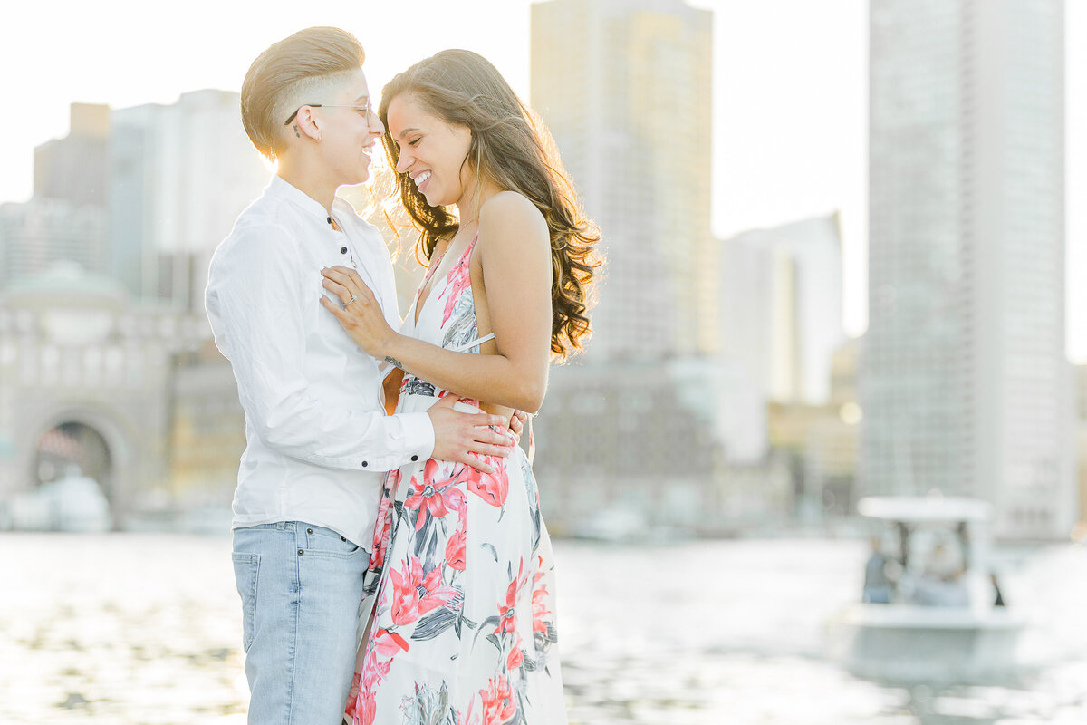 Couple are standing chest-to-chest and sharing an intimate laugh at their Boston Seaport Engagement Photoshoot. The city's skyline is featured in the background and boats passing by. Captured by New England Wedding Photographer Lia Rose Weddings